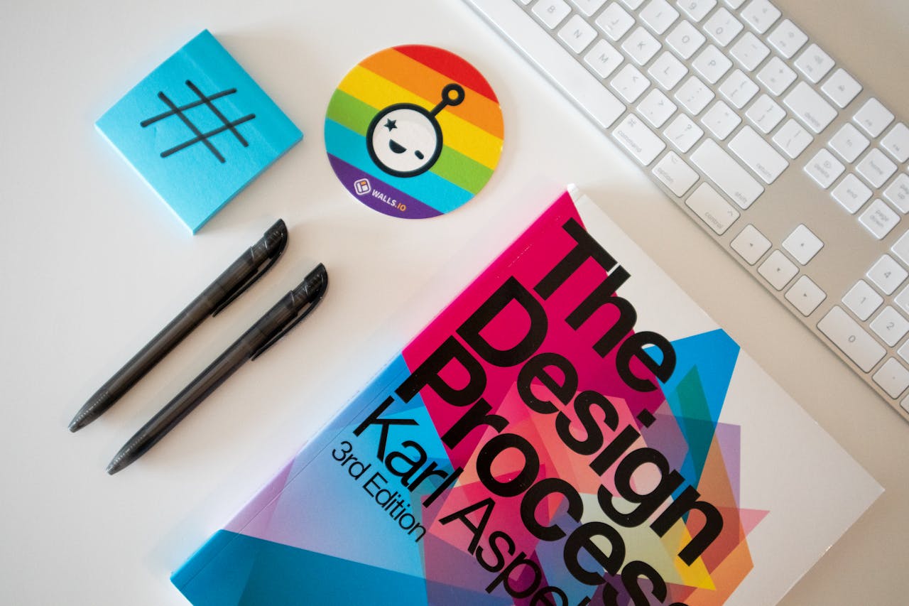 services-02 Flat lay of a creative desk setup featuring a design book, pens, and rainbow-themed sticker.