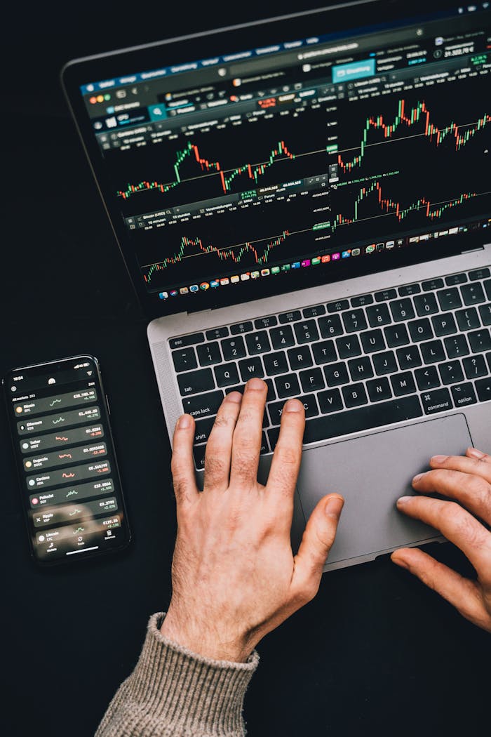 heros-img Hands typing on a laptop with cryptocurrency charts and smartphone display in view, focusing on digital finance.
